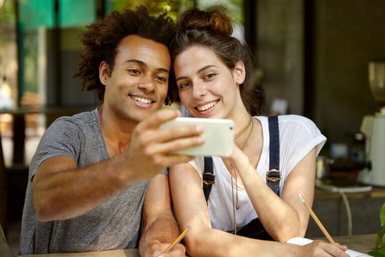 Lovely mixed race couple making selfie while sitting at cafeteria smiling broadly in camera of moder